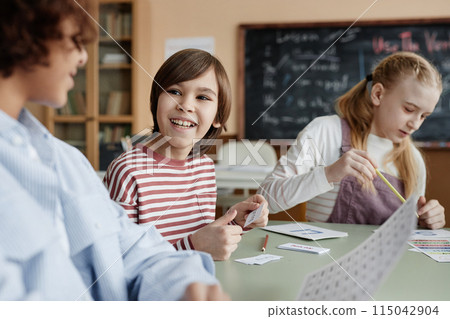 Cheerful Caucasian boy sitting at table in classroom chatting with his classmate during lesson Cheerful Caucasian boy sitting at table in classroom chatting with his classmate during lesson 115042904