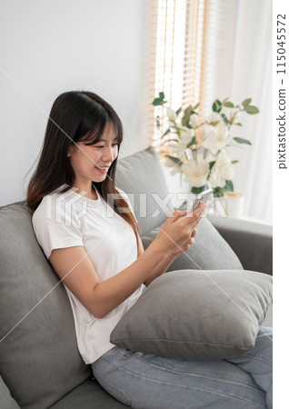 A young, happy Asian woman is relaxing on a sofa in the living room and using her smartphone. A young, happy Asian woman is relaxing on a sofa in the living room and using her smartphone. 115045572
