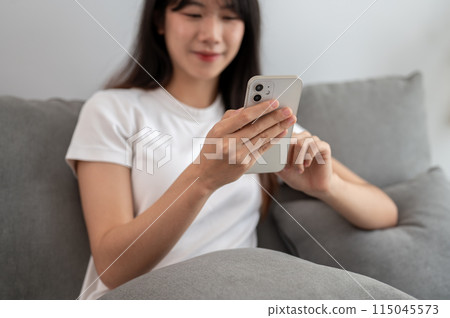 A young, happy Asian woman is relaxing on a sofa in the living room and using her smartphone. A young, happy Asian woman is relaxing on a sofa in the living room and using her smartphone. 115045573