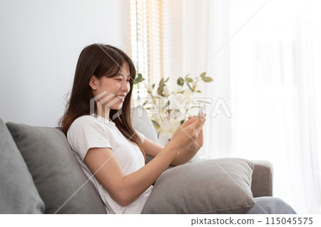 A young, happy Asian woman is relaxing on a sofa in the living room and using her smartphone. A young, happy Asian woman is relaxing on a sofa in the living room and using her smartphone. 115045575