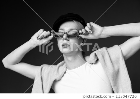 Black and white photo of young man with make up on his face wearing sunglasses. Man wearing fashionable sweater and cap. Man posing for a photo. Black and white photo of young man with make up on his face wearing sunglasses. Man wearing fashionable sweater and cap. Man posing for a photo. 115047726