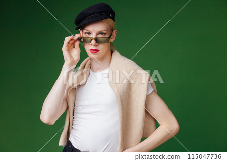 Young blond man with make up on his face and sunglasses. Man wearing fashionable white t-shirt and black cap. Man isolated on green background. Young blond man with make up on his face and sunglasses. Man wearing fashionable white t-shirt and black cap. Man isolated on green background. 115047736