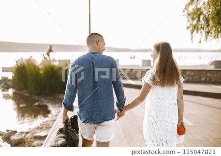 Romantic couple boyfriend and girlfriend walking with rotweiller dog near the lake together. Man and woman looking at each other. Man wearing blue shirt and woman white dress. 115047821