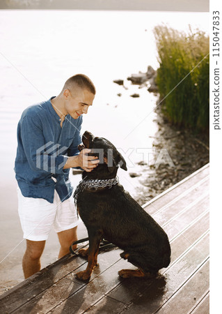 Handsome young male in casual outfit playing with cute dog while standing near the lake. Boy wearing blue shirt and white jeans shorts. Dog has a white and black scarf on his neck. 115047833