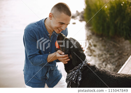 Handsome young male in casual outfit playing with cute dog while standing near the lake. Boy wearing blue shirt and white jeans shorts. Dog has a white and black scarf on his neck. Handsome young male in casual outfit playing with cute dog while standing near the lake. Boy wearing blue shirt and white jeans shorts. Dog has a white and black scarf on his neck. 115047849