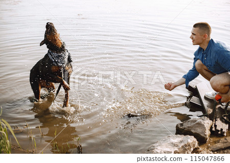 Handsome young male in casual outfit playing with cute dog while sitting near the lake. Boy wearing blue shirt and white jeans shorts. Dog has a white and black scarf on his neck. 115047866
