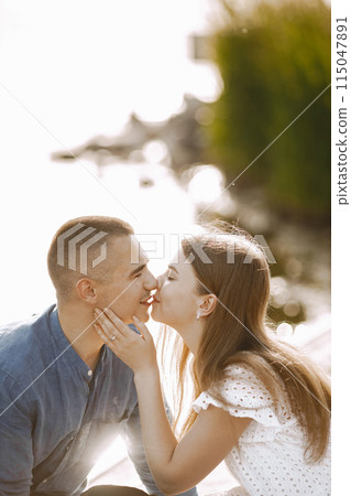 Romantic couple boyfriend and girlfriend kissing near the lake. Man and woman looking at each other and kissing. Man wearing blue shirt and woman white dress. Romantic couple boyfriend and girlfriend kissing near the lake. Man and woman looking at each other and kissing. Man wearing blue shirt and woman white dress. 115047891