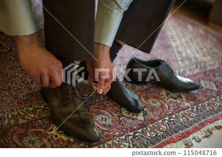 A man is tying leather dress shoes on an oriental rug in preparation for a formal event 115048716