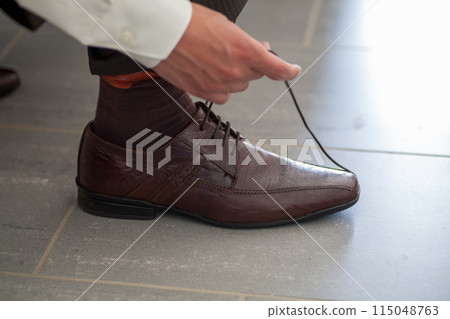 A man laces up a brown leather dress shoe on a grey tile floor, focusing on stylish accessories 115048763
