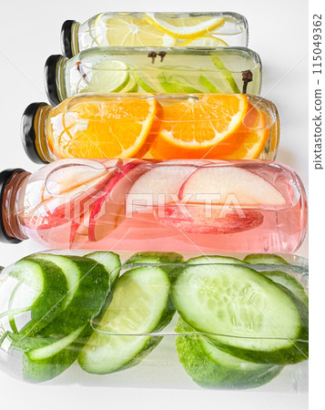 Stacked glass bottles with detox drinks with cucumber, apple, orange, and lemon slices on white background. Vertical orientation, refreshing beverage concept for design and print. Stacked glass bottles with detox drinks with cucumber, apple, orange, and lemon slices on white background. Vertical orientation, refreshing beverage concept for design and print. 115049362