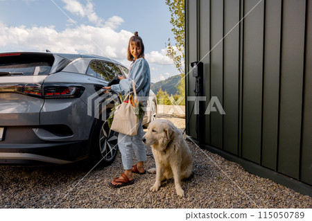 Woman plugging a charger into electric vehicle, with a dog near house 115050789