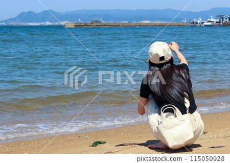 一個女人蹲在海邊的沙灘上，望著大海，一頭烏黑的長髮，戴著一頂白色的帽子。 115050928