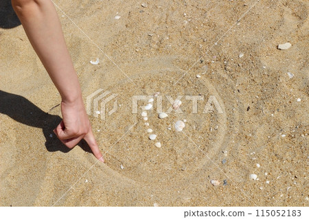 一位女士的手在沙灘上勾勒出一張由貝殼製成的臉的輪廓。 115052183