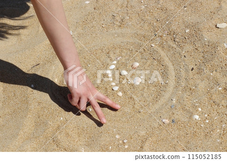 A woman's hand draws the outline of a face made with seashells on the beach with her fingers; her hand makes a peace sign A woman's hand draws the outline of a face made with seashells on the beach with her fingers; her hand makes a peace sign 115052185