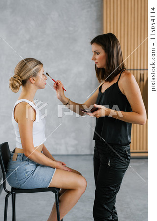 Makeup artist makes up a woman by applying powder with a brush. The woman is wearing a white T-shirt and blue jeans 115054114