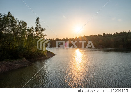 The sun sets over a calm lake, framed by trees in the foreground The sun sets over a calm lake, framed by trees in the foreground 115054135