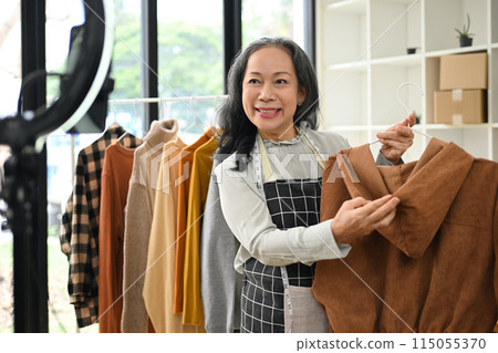 Attractive middle age small business owner live streaming and showing clothing to customer at her shop 115055370
