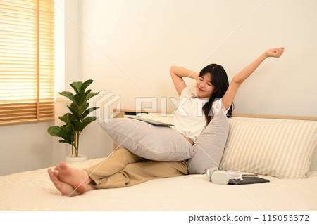 Carefree young Asian woman wearing casual clothes sitting in bed and stretching her arms Carefree young Asian woman wearing casual clothes sitting in bed and stretching her arms 115055372
