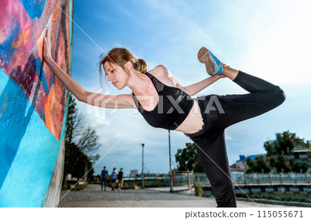 Young woman doing gymnastics on the street. Young woman doing sport exercise against colorful wall on city street. 115055671