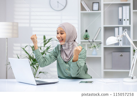 Happy woman in hijab enthusiastically celebrating achievement in front of a laptop in a bright, modern home office environment. Happy woman in hijab enthusiastically celebrating achievement in front of a laptop in a bright, modern home office environment. 115057258