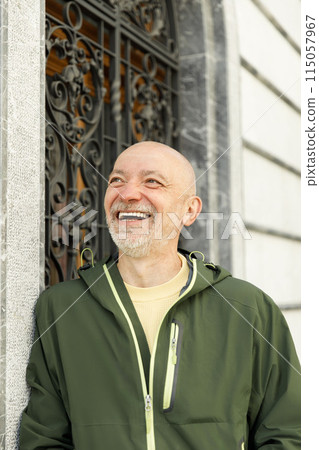 Content bald senior man with beard wearing a green Marmot jacket, smiling in a sunlit urban setting 115057967