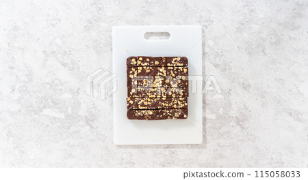 Flat lay. Cutting chocolate hazelnut fudge with a large kitchen knife into square pieces on a white cutting board. 115058033