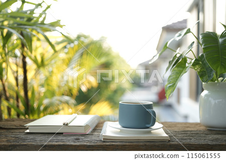 Blue coffee cup on wooden tray and notebook and Philodendron plant on wooden table under sunlight 115061555