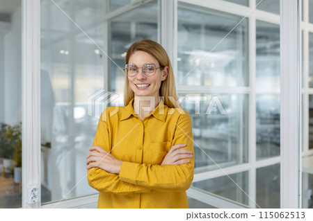 Confident businesswoman in a yellow shirt and glasses standing with arms crossed in a modern office setting. Confident businesswoman in a yellow shirt and glasses standing with arms crossed in a modern office setting. 115062513