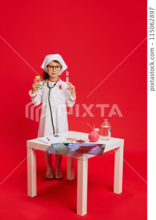 Little doctor. Child dressed in medical attire posing next to table with various medical supplies against vibrant red background. Little doctor. Child dressed in medical attire posing next to table with various medical supplies against vibrant red background. 115062897