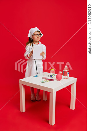 Little girl in doctor's costume, writing notes on folder standing by table of medical supplies against vibrant red background. 115062910