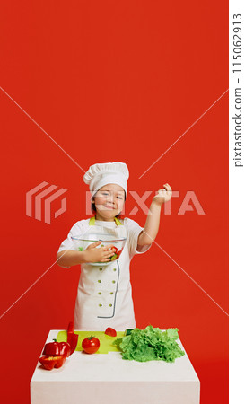 Little girl dressed as chef, holding bowl of fresh salad standing by table with vegetables, dreaming of cooking against red studio background. 115062913