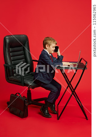 Kid CEO Making Deals. Young man dressed formal suit and talking on mobile phone against vibrant red studio background. Kid CEO Making Deals. Young man dressed formal suit and talking on mobile phone against vibrant red studio background. 115062965