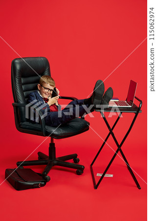 Young boy dressed in suit and tie sits confidently in office chair, talking on mobile phone with his feet up on table against vibrant red studio background. 115062974