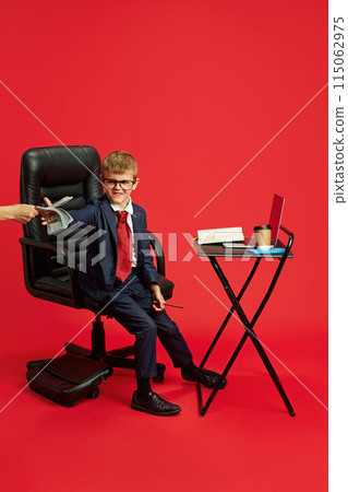 Boy dressed as businessman working in office and getting money against vibrant red studio background. 115062975