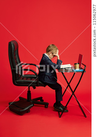 Portrait of little boy dressed as businessman sitting on office chair and working against vibrant red studio background. 115062977
