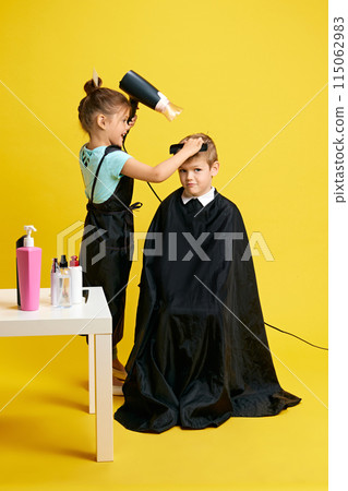 Cute little girl, acting as hairdresser, holds blowing and brushing hair of boy against vibrant yellow background. Cute little girl, acting as hairdresser, holds blowing and brushing hair of boy against vibrant yellow background. 115062983