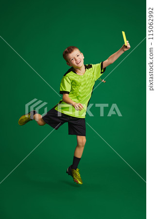 Little boy, kid referee showing yellow card and joyfully jumping against vibrant green background. Rules in soccer. 115062992