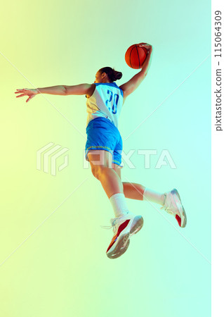 Low angle photo of athletic female basketball player leaping to make perfect shot in neon light against gradient studio background. 115064309