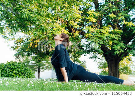 Relaxed middle aged plus size Caucasian woman performing the Upward Facing Dog yoga on a mat on green grass in park, exemplifying health and flexibility. Sport activity for wellbeing, mental health. 115064433
