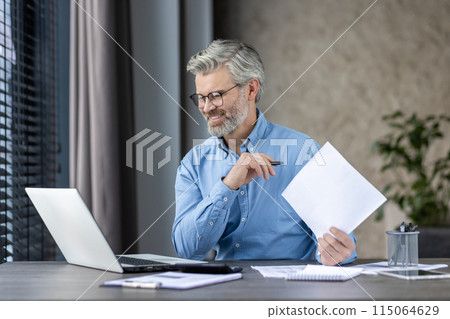 Smiling middle-aged man working at laptop home office hat home office holding papers and wearing glasses. 115064629