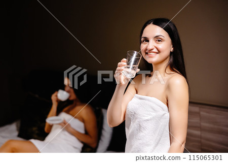 Cute young woman holding a glass of water in the morning while standing in the bedroom and behind her is her friend lying on the bed and drinking coffee 115065301