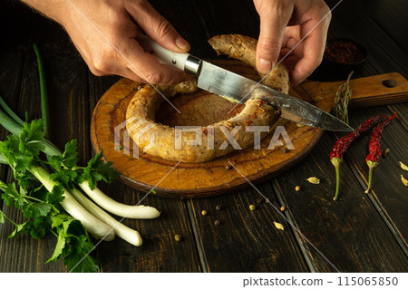 Close-up of a chef hands using a knife to cut meat sausage on a wooden cutting board. Cooking a national meat dish on a restaurant kitchen table Close-up of a chef hands using a knife to cut meat sausage on a wooden cutting board. Cooking a national meat dish on a restaurant kitchen table 115065850