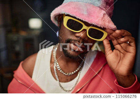 Close up portrait of Black young man wearing fuzzy bucket hat and trendy sunglasses looking at camera indoors Close up portrait of Black young man wearing fuzzy bucket hat and trendy sunglasses looking at camera indoors 115066276