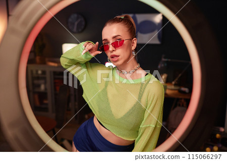 Waist up portrait of trendy young woman wearing neon outfit recording videos for social media shot through ring light 115066297