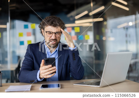 Upset and disappointed man with phone in hands at workplace, mature experienced businessman in business suit sad reading online message and checking email mail using app on smartphone. 115066526