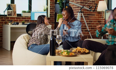Happy women chatting on cozy couch at apartment party, drinking wine and enjoying conversation. Cheerful friends catching up with each other at home in stylish living room, talking 115069078