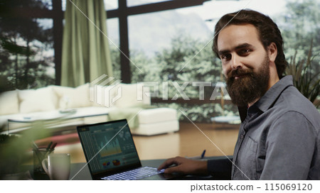 Young man sitting at desk in his luxury cabin with large windows showing panoramic forest view and opening pc, working on laptop thinking about new income development. Billionaire at mountain villa. Young man sitting at desk in his luxury cabin with large windows showing panoramic forest view and opening pc, working on laptop thinking about new income development. Billionaire at mountain villa. 115069120