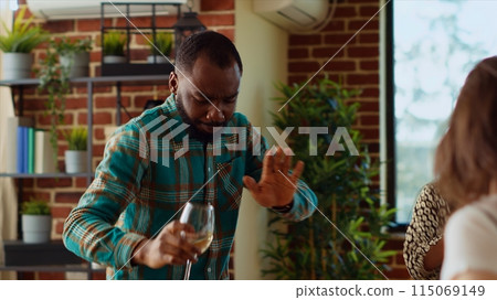 Guests at home party doing dance moves in brick wall living room, holding wine glasses in hand. African american man dancing with friends on energetic beats during weekend hangout, shaking hips Guests at home party doing dance moves in brick wall living room, holding wine glasses in hand. African american man dancing with friends on energetic beats during weekend hangout, shaking hips 115069149