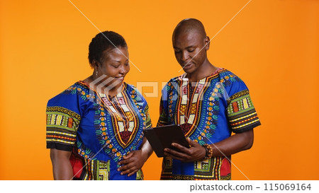 Ethnic people navigating on social media webpage, checking messages and looking at photos over orange background. African american man and woman scrolling online websites in studio. 115069164