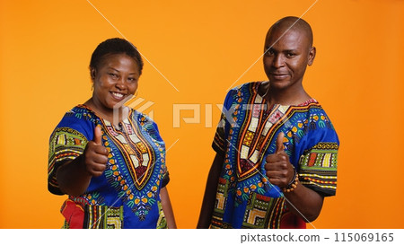 African american partners giving thumbs up gesture in studio, feeling cheerful and presenting their positive feedback. Married couple showing approval and doing like symbol on camera. 115069165